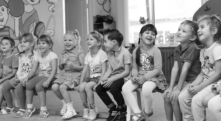 Eine Gruppe von Kindern sitzt freudig und lachend in einer Reihe auf Bänken vor einer Wand mit bunten Zeichnungen.  