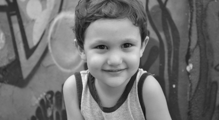 Foto eines kleinen Jungen mit dunkelbraunen kruzen Haaren. Er trägt ein graues Unterhemd und lächelt. Er steht vor einer bunten Wand mit blauen und grünen Grafitti-Motiven.