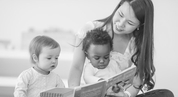 Eine Erzieherin sitzt mit 2 Kleinkindern auf dem Boden und schaut mti dem Kleinkindern gemeinsam ein Buch an. Ein Kleinkind sitzt auf dem Schoß der Erzieherin und das zweite Kind sitzt direkt links neben der Erzieherin..