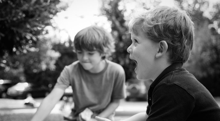 Zwei Jungen spielen im Freien und lachen. Im Hintergrund sind parkende Autos und Bäume zu erkennen.