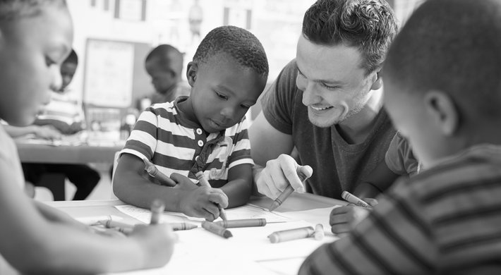 Bild zeigt eine Situation in einem Kindergarten. Ein Erzieher sitzt mit drei Kindern an einem Tisch. Die Kinder malen auf Papier.