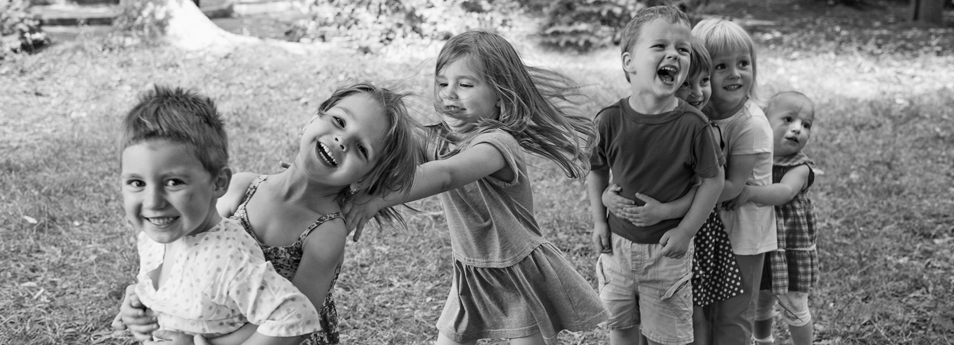 Sieben Kinder stehen in einer Reihe auf einer Wiese. Sie halten einander an den Schultern fest, ähnlich einer Polonaise. Sie lachen. Im Hintergund sind Bäume und Sträucher.