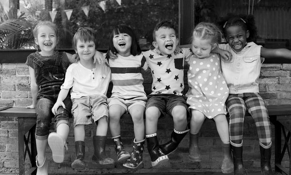 Sechs Kinder, Mädchen und Jungen, sitzen auf einer Bank für einer Backsteinwand. Die haben einander die Arme auf die Schultern gelegt und lachen in die Kamera. Alle tragen Gummistiefel.