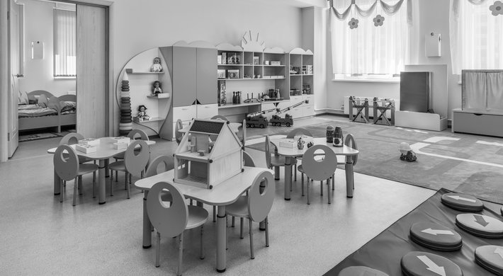 Das Bild zeigt einen Raum in einer Kita. Im Zentrum stehen drei Tische mit je vier Stühlen. Im Raum ist Spielzeug verteilt.
