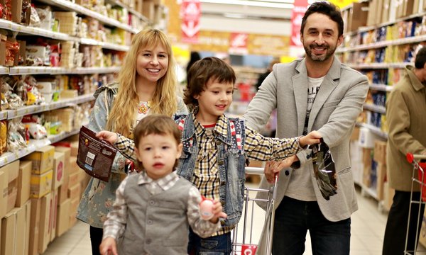 Foto zeigt Familie beim Einkaufen. Vater und MUtter schieben den Einkaufswagen. Die beiden Kinder sitzen im Einkaufswagen. Rechts und links von ihnen sind Supermarktregal zu sehen.