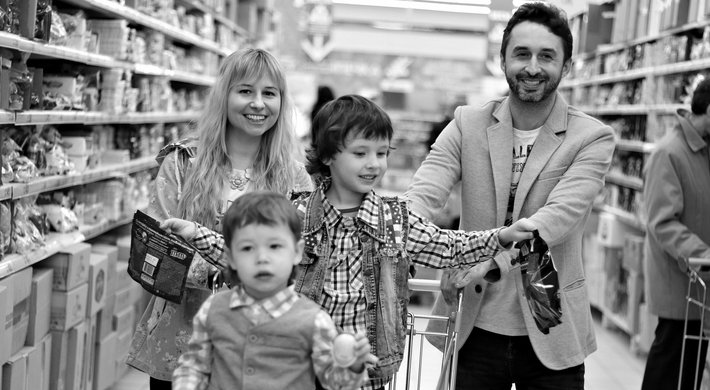 Foto zeigt Familie beim Einkaufen. Vater und MUtter schieben den Einkaufswagen. Die beiden Kinder sitzen im Einkaufswagen. Rechts und links von ihnen sind Supermarktregal zu sehen.
