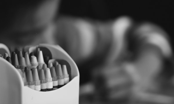Im Vordergrund steht eine gelbe Box mit bunten Wachsmalstiften. Hintergrund ist verschwommen und zeigt ein Kind, das malt.