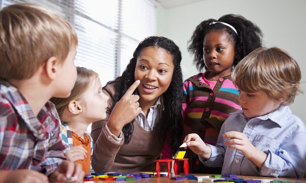 Erzieherin sitz mit vier Kindern um einen Tisch. Erzieherin erklärt den Kindern etwas und hält den Zeigefinder nach oben. Die KInder hören gespannt zu.