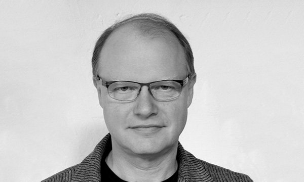 Schwarz-weiß Foto eines Mannes mit kurzen blonden Haaren. Er trägt ein graues Jacke und eine Brille mit schwarzem Oberrand.