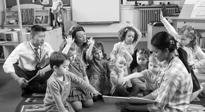 Foto in einem Kindergarten mit drei Erziehern, zwei davon sind Frauen. Zwischen ihnen sitzen acht kleine Kinder. Sie schauen gemeinsam in ein Buch. Einige Kinder heben die Hand. Im Hintergrund sind Spiezeuge zu erkennen.