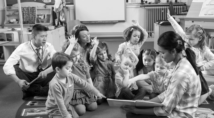 Foto in einem Kindergarten mit drei Erziehern, zwei davon sind Frauen. Zwischen ihnen sitzen acht kleine Kinder. Sie schauen gemeinsam in ein Buch. Einige Kinder heben die Hand. Im Hintergrund sind Spiezeuge zu erkennen.