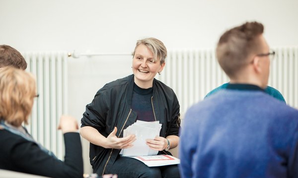 Das Bild zeigt Goska.Soluch sitzend mit Papier in den Händen vor einer Heizung. Einige Personen sitzen vor ihr. 