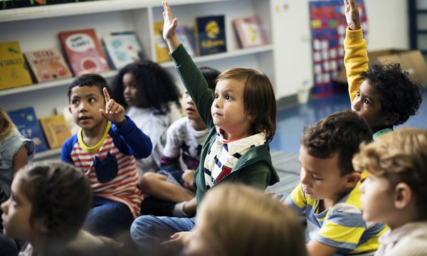 Kinder sitzen nebeneinander vor Regalen mit Broschüren. Einige von ihnen heben die Hand.