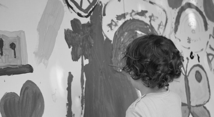 Kind, das mit dem Rücken zum Betrachter an eine Wand mit vielen bunten Motiven malt.