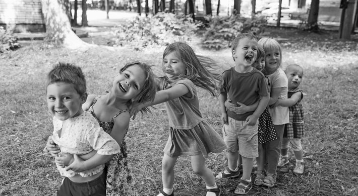 Sieben Kinder stehen in einer Reihe auf einer Wiese. Sie halten einander an den Schultern fest, ähnlich einer Polonaise. Sie lachen. Im Hintergund sind Bäume und Sträucher.