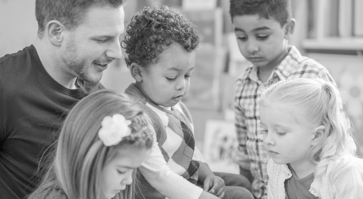 Männlicher Erzieher schaut sich gemeinsam mit vier Kindern ein Bilderbuch an.
